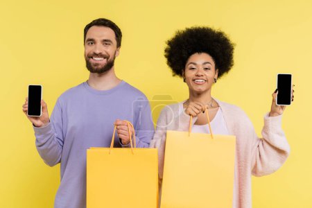 joyful multiethnic couple with shopping bags showing mobile phones with blank screen isolated on yellow