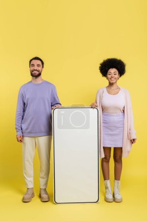 full length of joyful and stylish interracial couple standing near white template of cellphone on yellow background