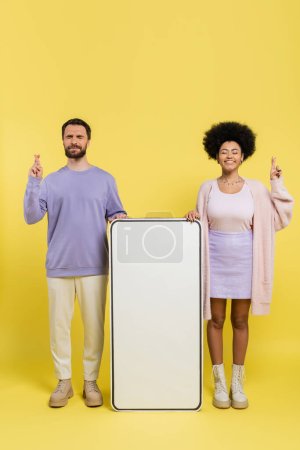 Photo for Full length of worried man and optimistic african american woman crossing fingers near mock-up of smartphone on yellow background - Royalty Free Image