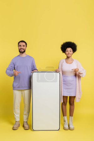 full length of cheerful multiethnic couple pointing at blank template of mobile phone on yellow background
