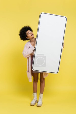 Photo for Full length of amazed african american woman holding huge cardboard model of smartphone on yellow background - Royalty Free Image