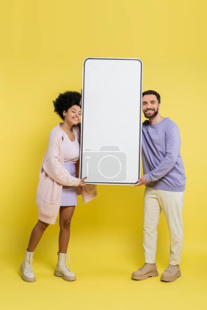 Photo for Full length of cheerful multiethnic couple holding blank template of smartphone on yellow background - Royalty Free Image