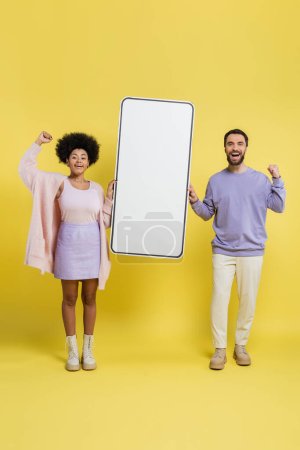 full length of excited interracial couple rejoicing near carton mock-up of mobile phone on yellow background