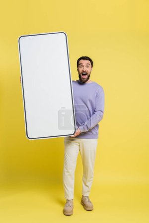 full length of astonished man with open mouth holding huge mock-up of smartphone on grey background