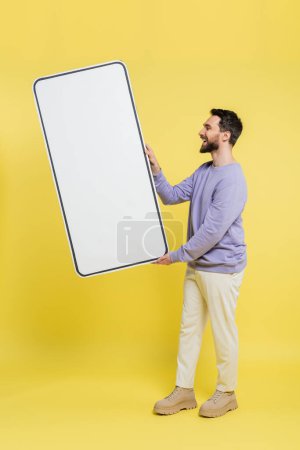 Photo for Side view of happy bearded man holding huge template of mobile phone on grey background - Royalty Free Image