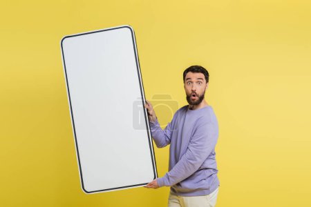 thrilled bearded man holding white template of mobile phone and looking at camera isolated on yellow