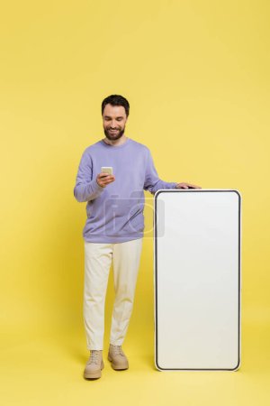 full length of positive man using smartphone near blank phone template on grey background