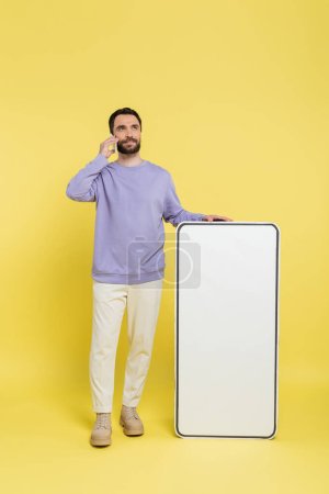 full length of bearded man talking on smartphone near huge phone template on grey background