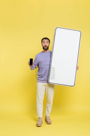full length of surprised man showing smartphone with blank screen near white template of mobile phone on grey background