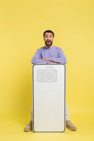 thrilled bearded man looking at camera near huge cardboard smartphone on yellow background