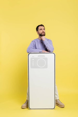 thoughtful bearded man looking away near mock-up of white mobile phone on yellow background