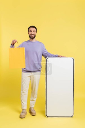 full length of cheerful bearded man standing with shopping bag and huge template of smartphone on grey background