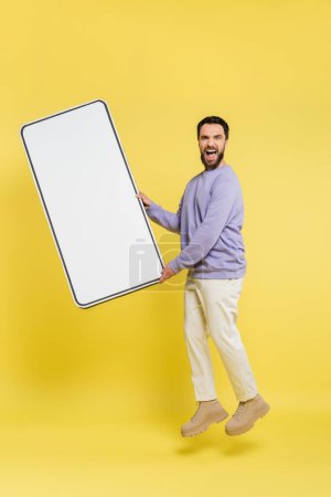 full length of excited bearded man levitating with huge mobile phone template on grey background