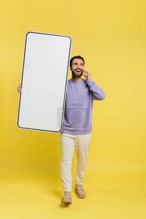 full length of happy man walking with huge phone mock-up and talking on cellphone on grey background