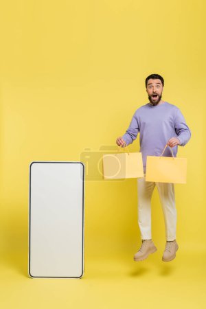 full length of excited man levitating with shopping bags near carton template of mobile phone on grey background