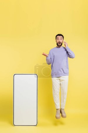amazed man talking on cellphone while levitating and pointing at huge mock-up of mobile phone on grey background