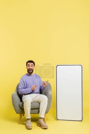 full length of astonished man sitting in armchair and pointing at big phone mockup on grey background
