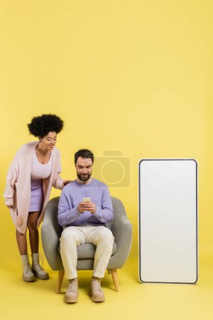 Photo for Full length of man using smartphone in armchair near smiling african american woman and huge phone template on yellow background - Royalty Free Image