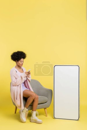 full length of stylish african american woman using mobile phone on armchair near white template of cellphone on yellow background