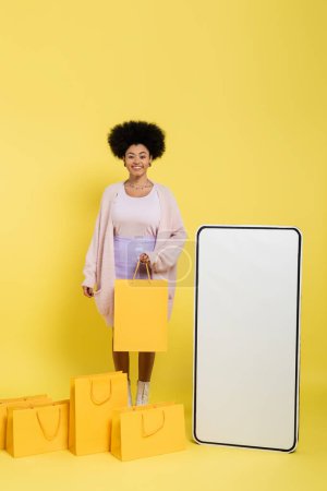 cheerful african american woman smiling at camera near shopping bags and white template of mobile phone on yellow 