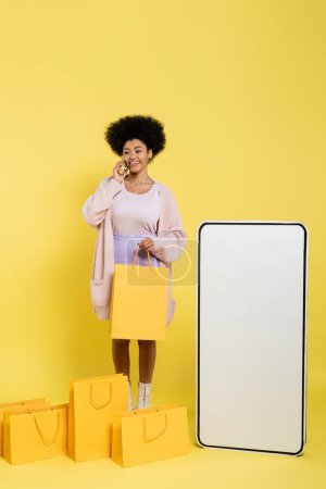 happy african american woman talking on smartphone near shopping bags and huge phone mock-up on yellow background
