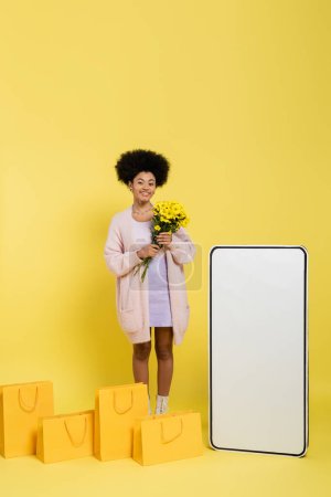 happy african american woman standing with flowers near shopping bags and blank template of smartphone on yellow background
