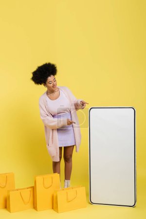 trendy african american woman pointing at huge template of mobile phone near shopping bags on yellow background