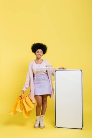 full length of joyful and trendy african american woman standing with shopping bags near carton model of smartphone on yellow background