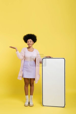 full length of cheerful african american woman pointing with hand near huge template of mobile phone on yellow background