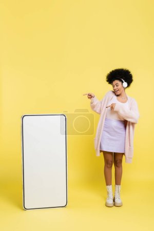 full length of happy african american woman in wireless headphones pointing at empty template of cellphone on yellow background