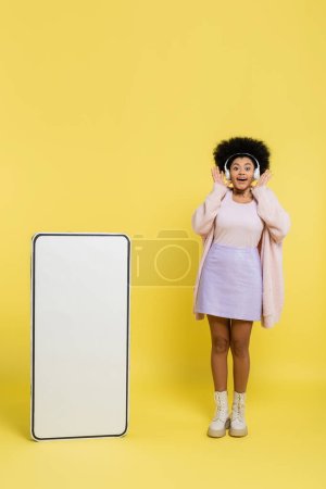 full length of excited african american woman in wireless headphones showing wow gesture near huge template of smartphone on yellow 