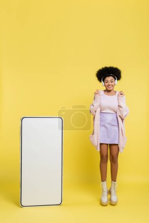 joyful african american woman in wireless headphones showing wow gesture while jumping near carton mobile phone on yellow 