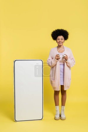 Photo for Full length of pleased african american woman standing with wireless headphones near white mock-up of cellphone on yellow background - Royalty Free Image