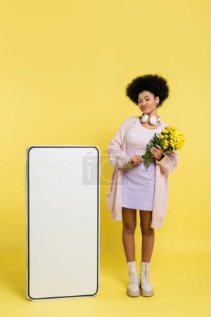full length of african american woman with wireless headphones and flowers smiling near blank template of smartphone on yellow 