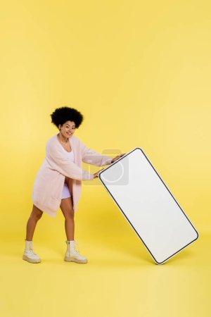 full length of smiling african american woman in cardigan and boots holding carton template of cellphone on yellow background