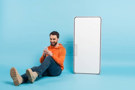 full length of smiling bearded man messaging on smartphone while sitting near huge phone template on blue background
