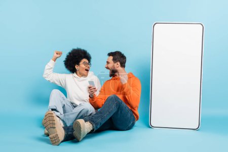 excited interracial couple showing win gesture while sitting near huge mobile phone mock-up on blue background