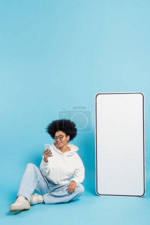 Photo for Full length of happy african american woman in eyeglasses sitting with smartphone near huge phone template on blue background - Royalty Free Image
