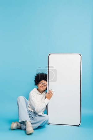 curly african american woman in eyeglasses and hoodie leaning on phone template while sitting with closed eyes on blue background