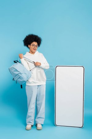 full length of smiling african american student with backpack looking at camera near empty phone template on blue background