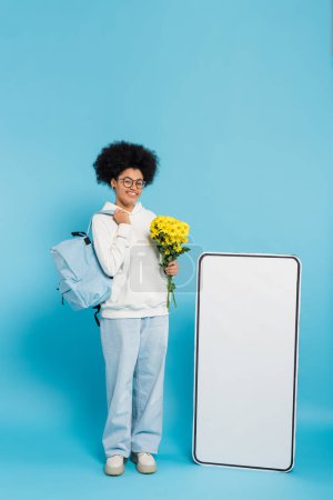 Photo for Full length of pleased african american student with yellow flowers and backpack near huge phone mock-up on blue background - Royalty Free Image