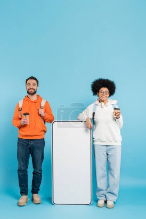 Foto de Happy interracial students with backpacks and coffee to go near white phone template on blue background - Imagen libre de derechos