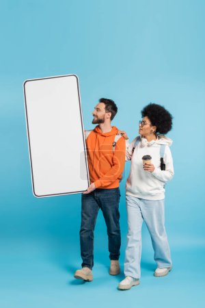 Téléchargez les photos : Bearded student carrying mobile phone template near african american girlfriend with coffee to go on blue background - en image libre de droit