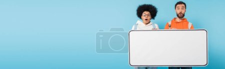 amazed interracial students looking at camera near huge phone mock-up isolated on blue, banner