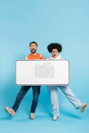 Foto de Full length of excited multiethnic students posing with big smartphone template and looking at camera on blue background - Imagen libre de derechos