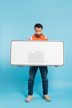 full length of astonished student holding white template of mobile phone on blue background