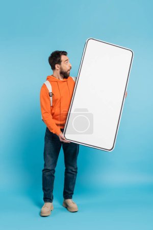 full length of amazed student in orange hoodie holding big phone template on blue background