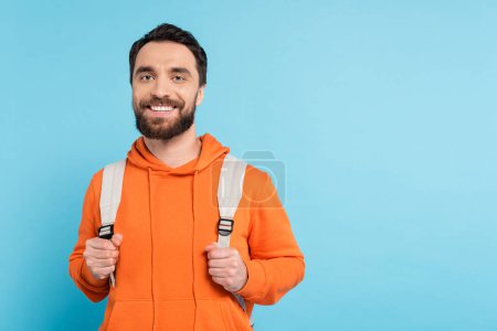 Foto de Happy bearded student in orange hoodie smiling at camera isolated on blue - Imagen libre de derechos