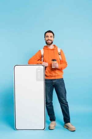 full length of bearded student with takeaway drink smiling at camera near white phone mock-up on blue background