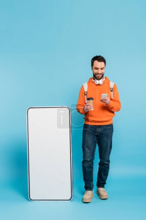 full length of smiling student with coffee to go using smartphone near carton mock-up of cellphone on blue background